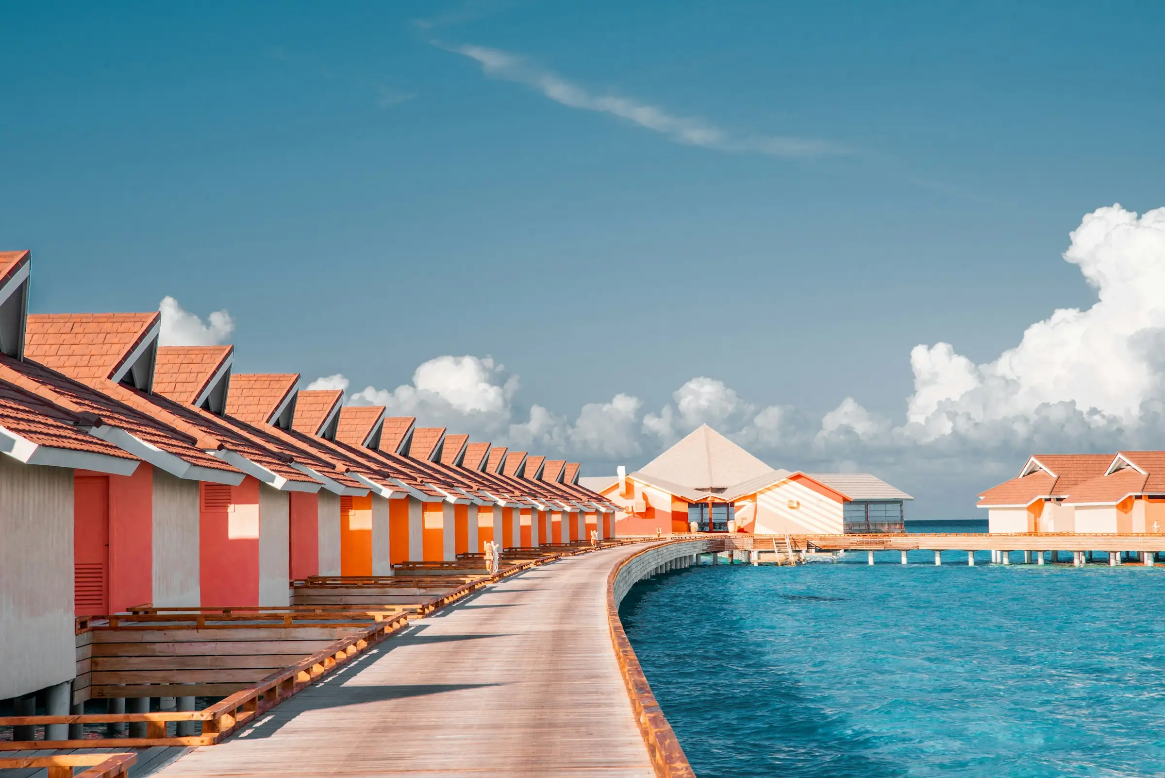 A wooden jetty curving past a row of overwater villas with orange roofs